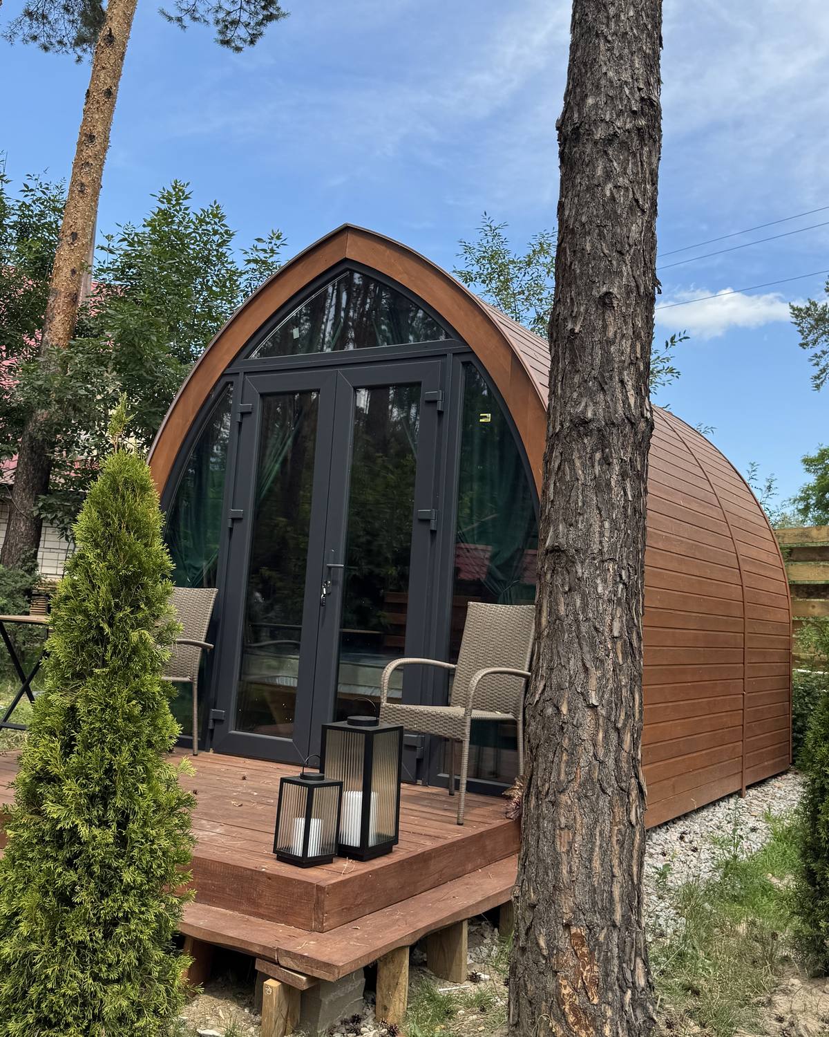 Збудований модульний будинок GoodWoodHome серед сосен: арковий фасад і тераса з кріслами та ліхтарями